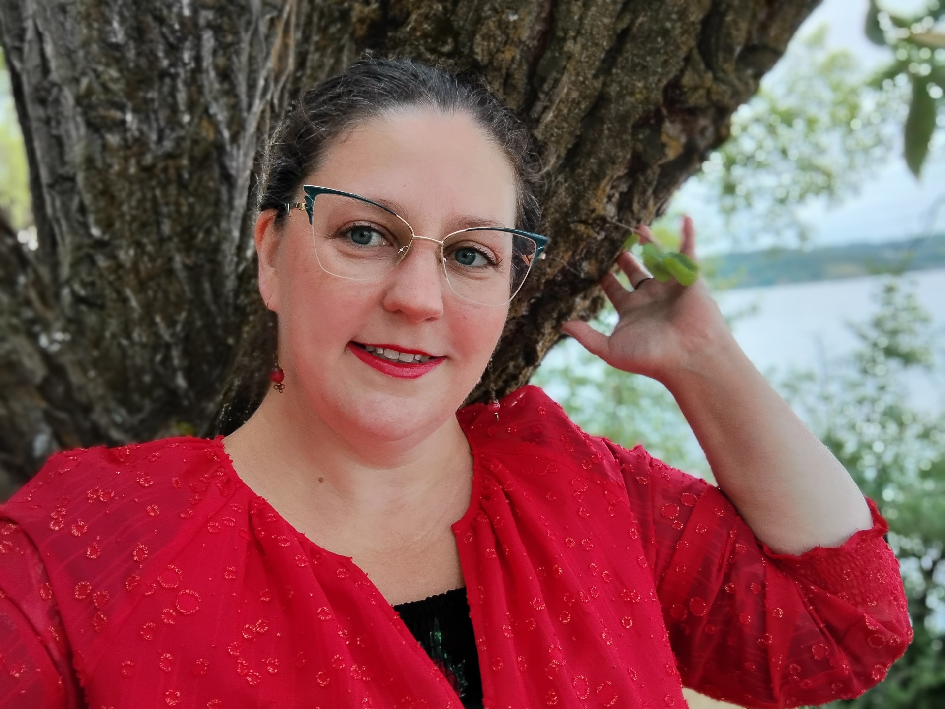 Brynna Lyon beside a tree by the water, wearing a red blouse